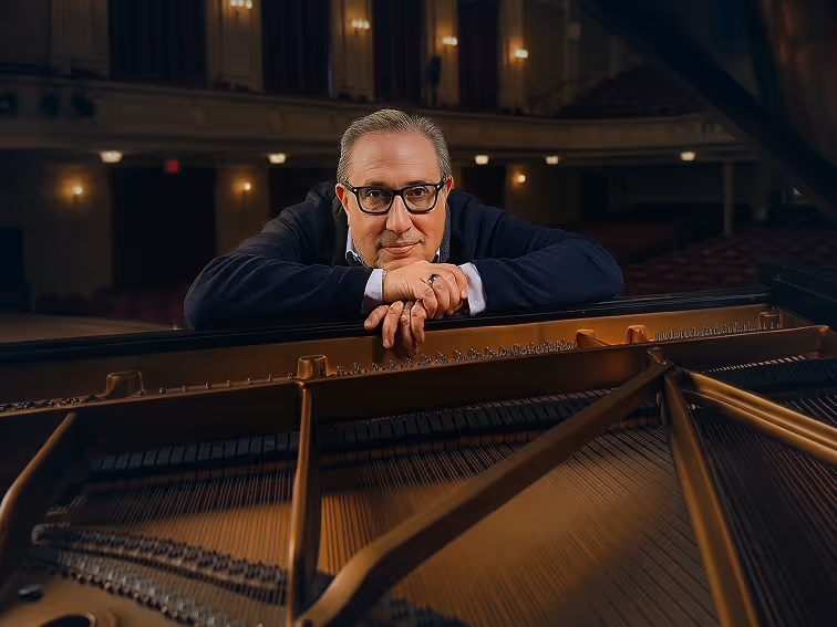 Man with glasses leaning on a grand piano inside a dimly lit concert hall.